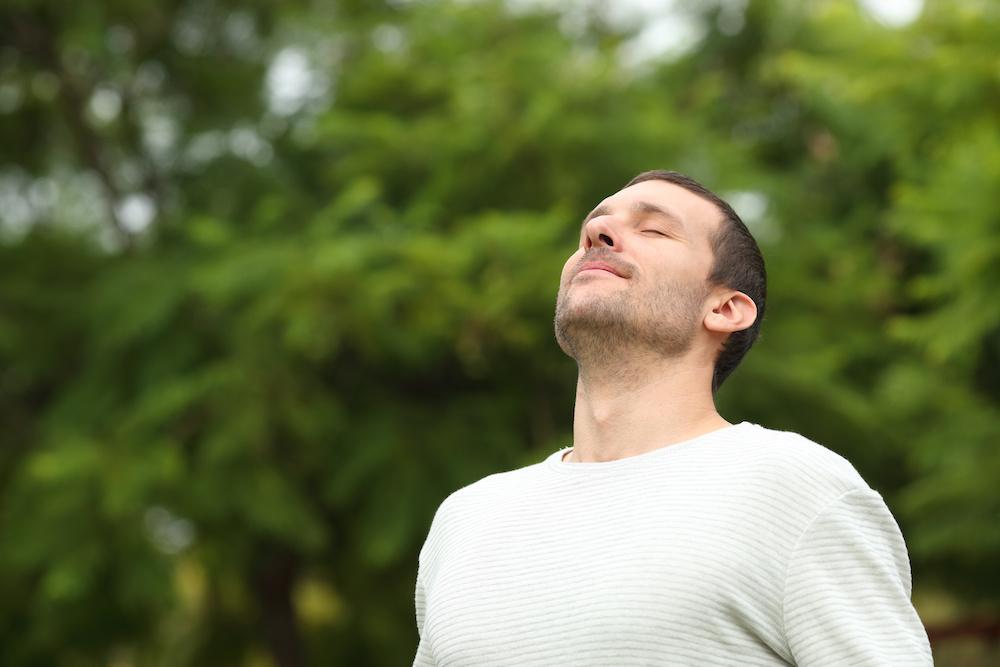 A man standing outdoors with his eyes closed, taking a calm breath